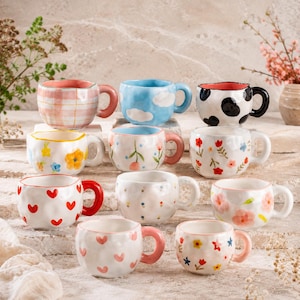 May include: A collection of ceramic mugs with various designs. Some mugs feature patterns like clouds, cow spots, and hearts. Others have floral and plaid designs. Each mug has a unique handle color, including red, pink, and black.