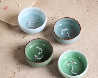 Könnte beinhalten: Vier kleine, runde Keramikschalen in Blau- und Grüntönen. Jede Schale zeigt ein detailliertes Design von Koi-Fischen, die darin schwimmen. Ideal zum Servieren von Tee oder kleinen Portionen.