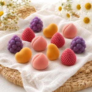 May include: A collection of small, fruit-shaped decorative items including purple blackberries, red strawberries with a textured surface, smooth yellow mangoes, and soft pink peaches, all arranged together on a white fabric cloth.
