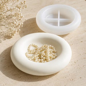 May include: A white circular tray with a smooth, rounded rim containing a strand of white faux pearls, positioned in front of a translucent silicone mould that features a circular shape with four internal dividers for casting.