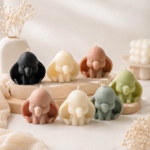 Peut inclure: Une collection de sept bougies en forme d'éléphant dans des couleurs variées, notamment noir, crème, beige, vieux rose et vert sauge. Chaque bougie présente une mèche centrale, de grandes oreilles et une trompe, disposées sur un support en bois.