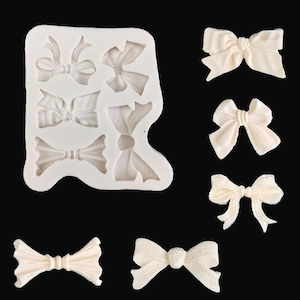 May include: A white silicone mould featuring five distinct recessed bow shapes, accompanied by five individual off-white bow-shaped castings arranged to the right and bottom of the mould, all displayed against a solid black background.