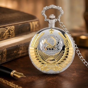 Puede incluir: Reloj de bolsillo plateado y dorado con cadena. La esfera del reloj presenta un emblema dorado con una hoz y un martillo sobre un globo, rodeado de espigas de trigo. El reloj tiene una parte superior plateada decorativa y una cadena.