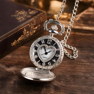 May include: A silver-toned pocket watch on a matching chain, featuring an ornate circular case with a heart-shaped inner dial. The white face displays black hands, black Arabic numerals, and the word QUARTZ printed in black on the dial.