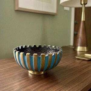 May include: A round bowl featuring a textured dark interior and an exterior with alternating vertical stripes of metallic gold and bright blue. The rim of the bowl has a notched, crenelated design, and the base is a solid, circular metallic gold pedestal.