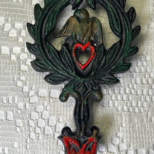May include: A dark metal decorative object featuring a central eagle figure enclosed within a green-painted laurel wreath. A bright red heart shape is positioned below the eagle, and a red decorative element is at the base of the handle.