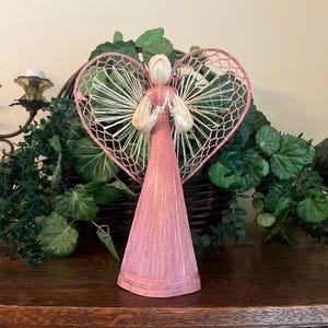 May include: A decorative angel figurine crafted from natural straw fibers. The figure features a long, conical pink dress, a light beige head, and large, heart-shaped wings constructed from pink-dyed straw mesh with radiating straw spokes.