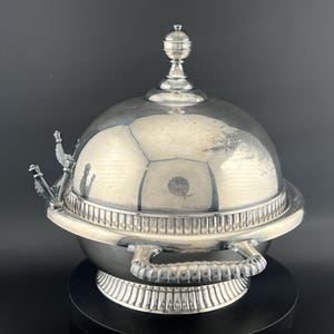 May include: A silver-colored metal serving dish with a domed lid, a spherical finial, and two side handles. The base and the lid rim feature a vertical fluted pattern. Two small decorative metal arrow-shaped implements are attached to the side.