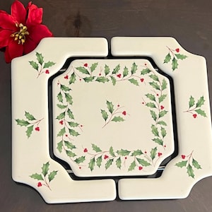 May include: An expandable ceramic trivet featuring a cream-colored surface decorated with green holly leaves and red berries. The square-shaped center piece is surrounded by four corner sections, all adorned with a matching holly and berry pattern.