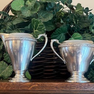 May include: A silver-toned metal creamer pitcher and a matching sugar bowl set. Both pieces feature a smooth, polished finish, a pedestal base, and a decorative embossed band around the upper rim. The creamer has a pouring spout and one handle.