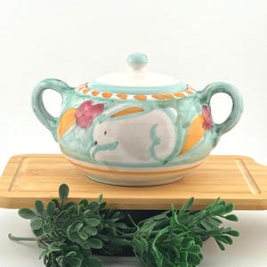 May include: A ceramic sugar bowl with a lid, featuring two side handles and a hand-painted design of a white rabbit, orange accents, and red floral motifs on a light green and white background, finished with a decorative orange dotted border.