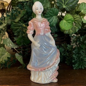 May include: A porcelain figurine of a person standing in a long dress with a pink bodice and a light blue skirt. The figure has white hair, painted facial features, and gold-colored trim accents along the sleeves, neckline, and hem of the dress.