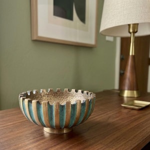 May include: A round metal bowl featuring a textured, hammered interior surface and a exterior design of alternating vertical brass-toned panels and recessed teal-colored grooves, finished with a notched, gear-like rim and a circular pedestal base.