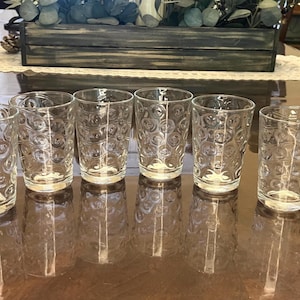 May include: A set of nine clear glass tumblers arranged in a row, each featuring a repeating embossed circular pattern across the exterior surface. The glasses are cylindrical with a flat base and a smooth rim, reflecting light on their transparent walls.