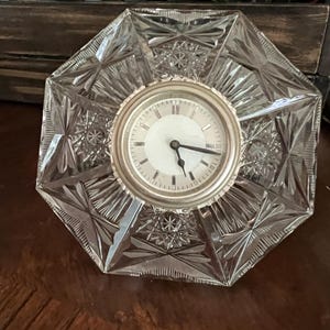 May include: An octagonal clear glass clock featuring a central circular face with a silver-toned bezel, black hands, and a white dial. The glass body is decorated with intricate geometric starburst patterns and faceted edges throughout the frame.