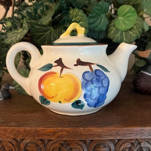 May include: Cream-colored ceramic teapot with a yellow handle and lid. The teapot is decorated with a large yellow apple and a cluster of blue grapes. Green leaves and brown stems accent the fruit design. The teapot sits on a wooden surface.