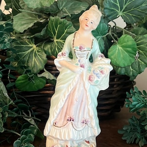 May include: A porcelain figurine of a person wearing a pale green and pink gown with ruffled edges and gold trim. The figure holds a basket of small painted flowers and stands on a circular base decorated with floral accents and a textured pattern.