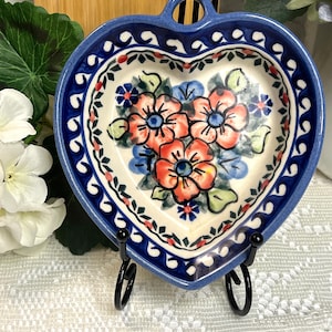 May include: A heart-shaped ceramic dish featuring a cream-colored center with a hand-painted floral pattern of orange and blue flowers. The rim is decorated with a dark blue border and small red accents. The dish includes a small hole at the top.