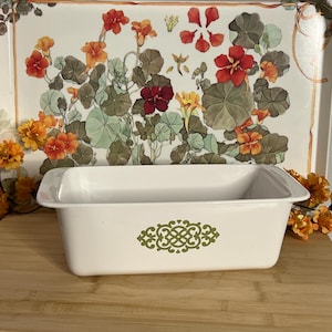 May include: A rectangular white ceramic loaf pan featuring a centered olive green filigree pattern on the front side. The bakeware has smooth, rounded edges and a deep interior basin, presented as a single functional kitchen vessel for baking.