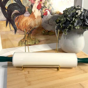 May include: A kitchen rolling pin featuring a smooth, cylindrical white barrel and two dark green handles on either side. The rolling pin is positioned horizontally on a gold-toned metal stand, showcasing its clean lines and contrasting color scheme.