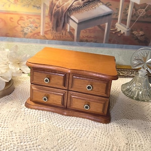 May include: A wooden jewelry chest with a smooth, light brown finish featuring four rectangular drawers arranged in two rows. Each drawer is equipped with a small, round, dark-colored knob with a decorative, light-colored scalloped edge.