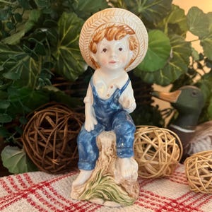 May include: A porcelain figurine of a young person wearing a straw hat, a white collared shirt, and blue overalls. The figure is seated on a textured tree stump base with green painted details, featuring a glossy finish and hand-painted facial features.