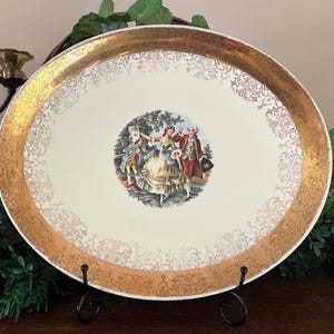 May include: An oval ceramic serving platter with a cream-colored center featuring a colorful illustration of two figures in 18th-century attire. The wide rim is decorated with a metallic gold border and an intricate, repeating filigree pattern.