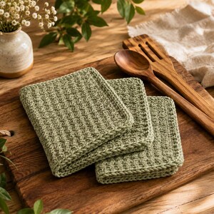 May include: Three square sage green crochet dishcloths with a textured waffle stitch pattern and a thick braided border. The dishcloths are stacked neatly, showcasing the consistent yarn tension and the soft, muted green color of the cotton material.