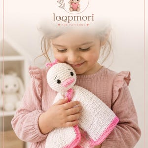 May include: A white and pink crochet security blanket featuring a round duck head with black eyes and a pink beak. The blanket has a textured white center with a pink border. The text loopmori PDF PATTERNS and CROCHET WITH LOVE is visible.