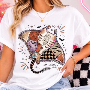 Könnte beinhalten: Ein weißes Kurzarm-T-Shirt mit einer großen Grafik eines Stachelrochens im Patchwork-Design. Das Rochen-Muster enthält Kürbisse, Geister, Fledermäuse, Spinnennetze und einen schwarz-weiß karierten Bereich, akzentuiert durch kleine Sterne und Wellen.