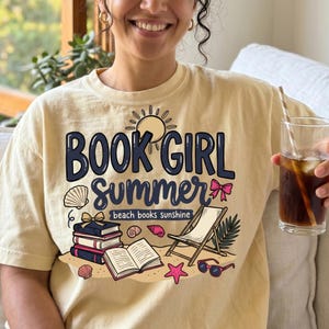 Puede incluir: Una camiseta de color crema con un estampado gráfico que incluye el texto "BOOK GIRL SUMMER" y "beach books sunshine". El diseño muestra un sol, una pila de libros, un libro abierto, una silla de playa, conchas, una estrella de mar y gafas de sol.