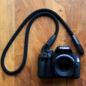 Pode incluir: Câmera DSLR Canon EOS 550D preta com uma alça de pescoço preta, em uma superfície de madeira. A lente da câmera está centralizada e o logotipo da Canon é visível. A alça é feita de material tecido.