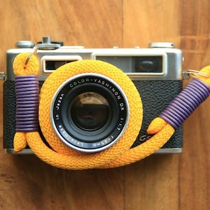 Op de afbeelding: Een vintage zilveren en zwarte filmcamera met een lens voorzien van de tekst COLOR-YASHINON DX 1:1.7 f=45mm. Een felgele gevlochten touwriem met paarse koordaccenten is rond de lens gewikkeld en aan de zijkanten van de camerabehuizing bevestigd.