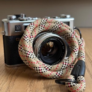 Könnte beinhalten: Eine silber-schwarze Vintage-Filmkamera mit einem Color-Yashinon DX 1:1.7 45mm Objektiv, teilweise umwickelt mit einem gewickelten Kameragurt. Der Gurt hat ein gewebtes Muster in Beige, Schwarz, Weiß und Rot und schwarze Kunststoffanschlüsse.