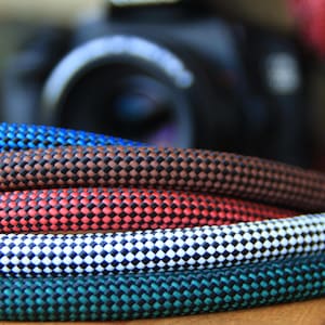 Puede incluir: Primer plano de varias correas de cámara coloridas. Las correas están hechas de un material tejido en varios colores, incluyendo azul, marrón, rojo, blanco y verde azulado. La cámara está borrosa en el fondo.