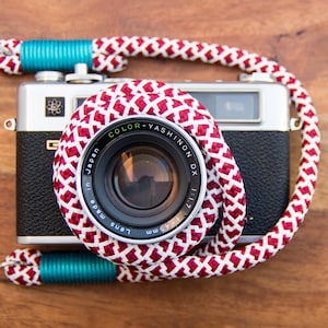 Könnte beinhalten: Eine silberne und schwarze Vintage-Kamera mit einem rot-weiß gemusterten Gurt. Das Objektiv ist mit "COLOR-YASHINON DX 1:1.7" und "45mm" beschriftet. Der Kameragurt hat türkisfarbene Akzente.