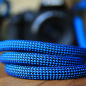 Op de afbeelding: Close-up van een opgerold blauw en zwart touw, waarschijnlijk voor klimmen. Het touw heeft een getextureerd patroon en is scherp gefocust, met een wazige achtergrond. De kleur is helderblauw met zwarte accenten.