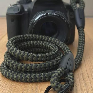 Könnte beinhalten: Schwarze Canon DSLR-Kamera mit einem Canon EF 50mm 1:1.8 Objektiv. Ein grün-schwarzer geflochtener Kameragurt ist vor der Kamera aufgerollt. Der Gurt hat einen schwarzen Anschluss und einen schwarzen Metallring.