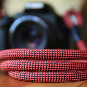 May include: A coiled red and black camera strap. The strap is made of a woven material with a repeating pattern of red and black dots. A black camera is slightly out of focus in the background.
