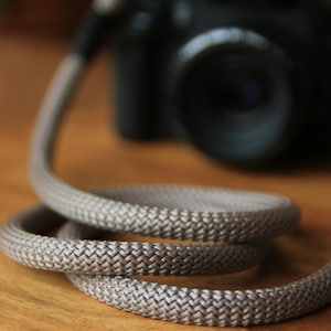 May include: A coiled, woven camera strap in a neutral beige colour. The strap is made of a thick, durable material and is designed to be worn around the neck. A black camera body is visible in the background, resting on a wooden surface.