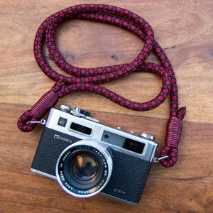 May include: A vintage Yashica GSN camera with a red and black patterned neck strap. The camera lens is labelled "Color-Yashinon DX 1:1.7 f=45mm". The strap is made of a thick, woven material.