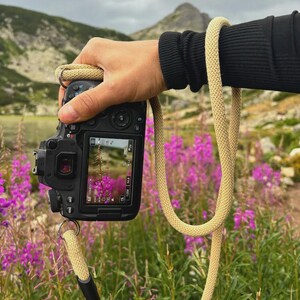 May include: A black digital camera with a beige rope strap is held in front of a field of purple flowers. The camera's screen displays a close-up of a purple flower. The background features a mountain and a cloudy sky.