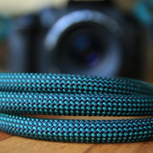 May include: A coiled teal and black camera strap with a woven design. The strap is in focus, with a blurred camera body behind it. The strap is made of a durable, textured material.