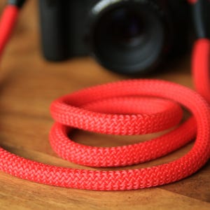 Op de afbeelding: Een felrode camerariem opgerold op een houten oppervlak. De riem is gemaakt van een dik, geweven materiaal en is bevestigd aan een zwarte camerabody. Het levendige rood van de riem contrasteert met de camera, wat een aantrekkelijk accessoire creëert.