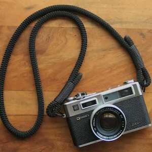 May include: A classic Yashica GSN 35mm film camera with a black and silver body and a black neck strap. The camera lens is labelled "Color-Yashinon DX 1:1.7 f=45mm". The strap is made of a thick, black woven material.