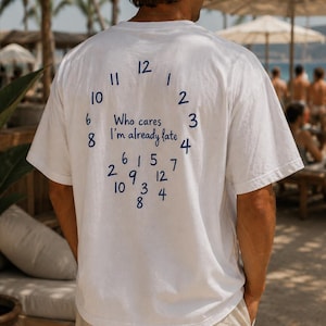 May include: A white short-sleeved t-shirt featuring a blue graphic on the back. The design displays numbers arranged in a circular clock pattern with the text "Who cares I'm already late" in the center, followed by additional scattered numbers.