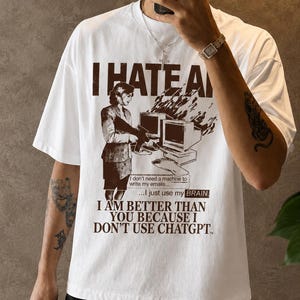 Peut inclure: Un t-shirt blanc à manches courtes avec une impression graphique marron représentant une personne debout près d'un ordinateur. Le texte indique : I HATE AI, I don't need a machine to write my emails... I just use my BRAIN. I AM BETTER THAN YOU BECAUSE I DON'T USE CHATGPT.