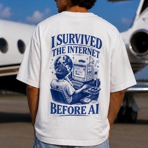 Könnte beinhalten: Ein weißes T-Shirt mit einer blauen Grafik auf der Rückseite. Das Design zeigt eine Person, die vor einem Computer sitzt, mit einem Globus, einem Mauszeiger und dem Text I SURVIVED THE INTERNET BEFORE AI. Das Shirt hat kurze Ärmel und einen Rundhalsausschnitt.