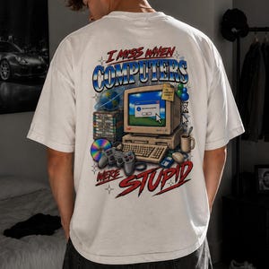 May include: A white t-shirt featuring a graphic print on the back. The design displays a vintage beige computer, a keyboard, a game controller, a stack of software boxes, a CD, and a mug. The text reads I MISS WHEN COMPUTERS WERE STUPID.