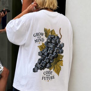 Könnte beinhalten: Ein weißes T-Shirt mit kurzen Ärmeln und einem Grafikdruck auf der Rückseite. Das Motiv zeigt eine Weintraube aus schwarzen Computertastaturtasten, umgeben von grünen Blättern. Der Text lautet GROW YOUR MIND und CODE YOUR FUTURE.